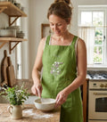 A woman wearing a green embroidered linen apron, whisking a bowl in a kitchen — hand-embroidered by The Thread Nest