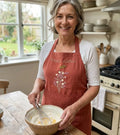 A woman wearing a rust linen apron embroidered with flowers and a kitchen name — hand-embroidered by The Thread Nest