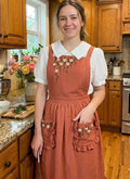A woman wearing a rust ruffled linen apron with white floral embroidery in a kitchen — hand-embroidered by The Thread Nest