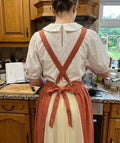 Back view of a woman wearing a rust ruffled apron with crossed straps tied in a bow — hand-embroidered by The Thread Nest
