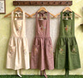 Three embroidered linen aprons in cream, mauve and green hanging on wooden hangers — hand-embroidered by The Thread Nest