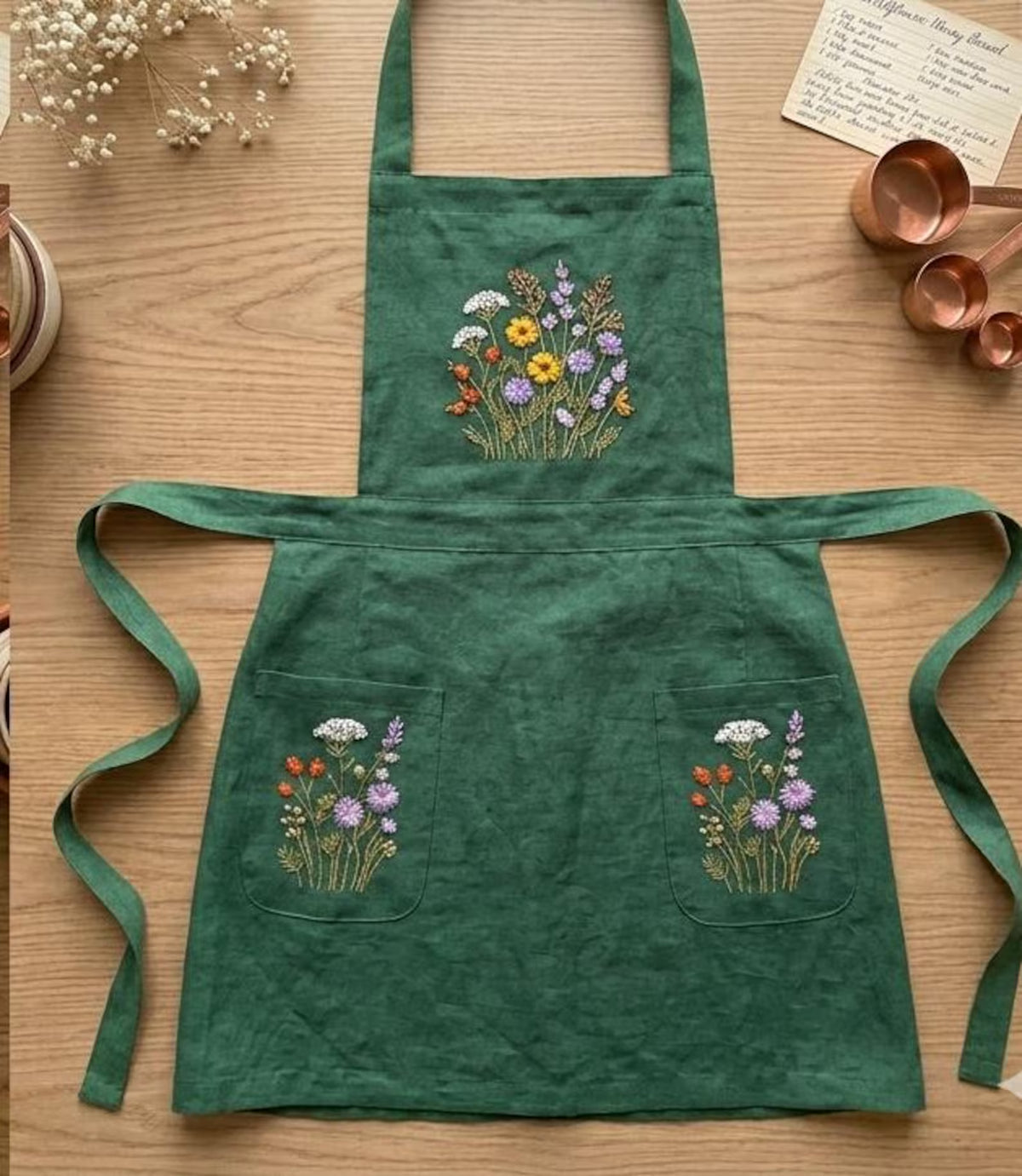 A dark green linen apron laid flat with an embroidered wildflower bouquet and two pockets — hand-embroidered by The Thread Nest