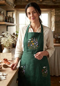 A woman wearing a dark green linen apron with embroidered wildflowers in a kitchen — hand-embroidered by The Thread Nest