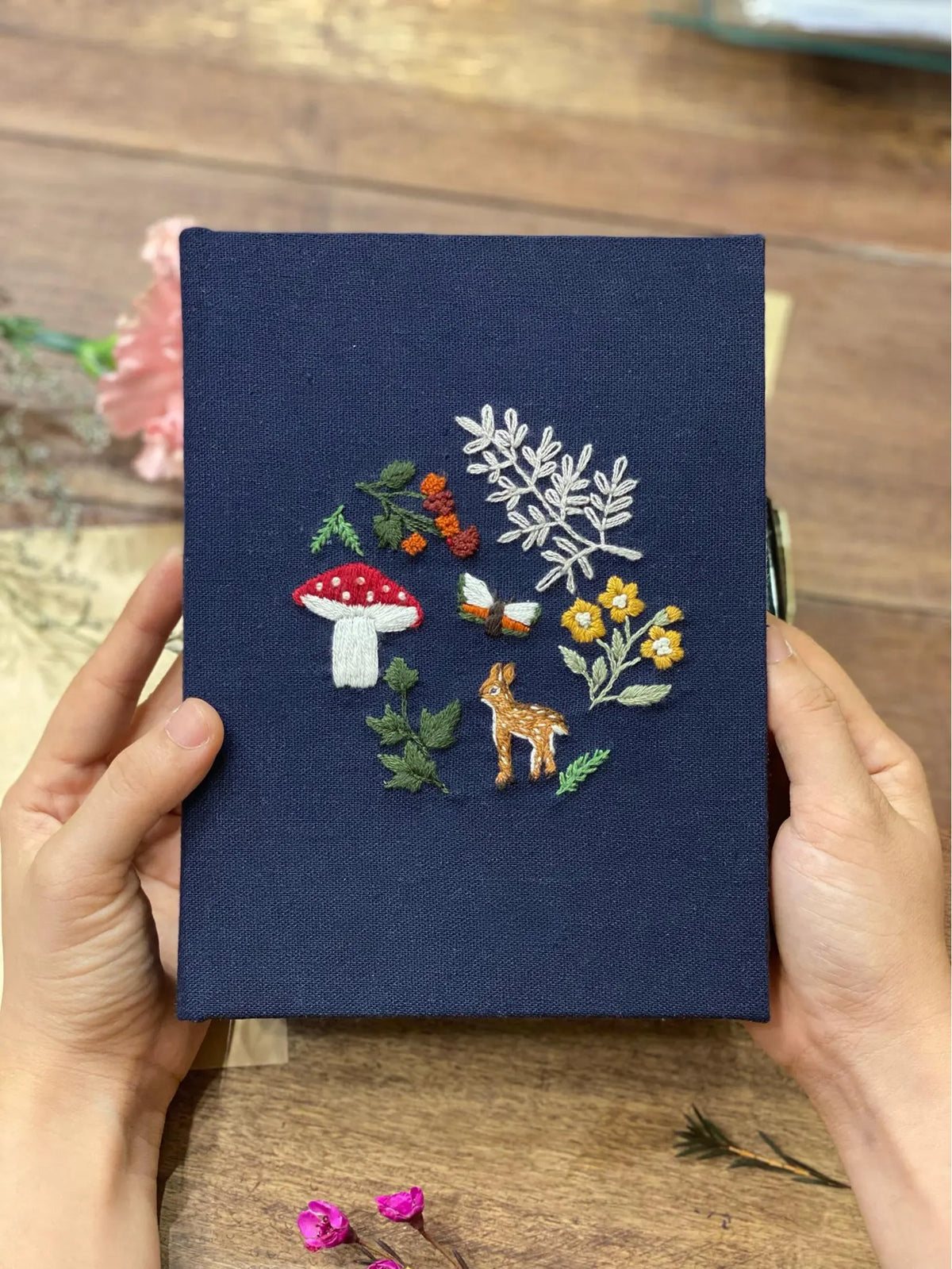 A navy blue journal with an embroidered mushroom, butterfly, deer and flowers — hand-embroidered by The Thread Nest