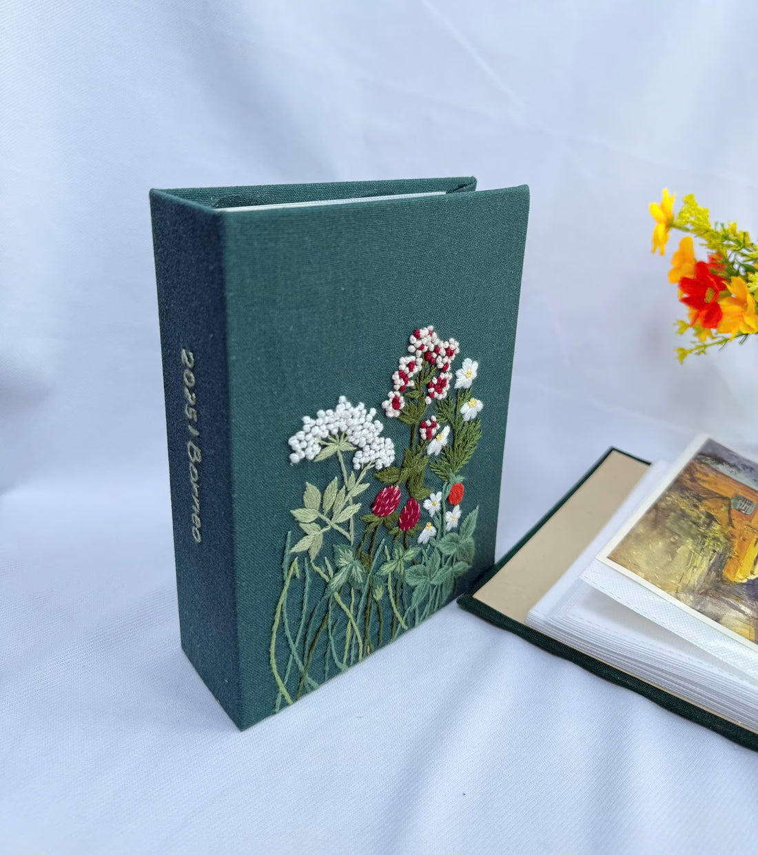 A dark green fabric-covered photo album with an embroidered floral design in white, red and green — hand-embroidered by The Thread Nest