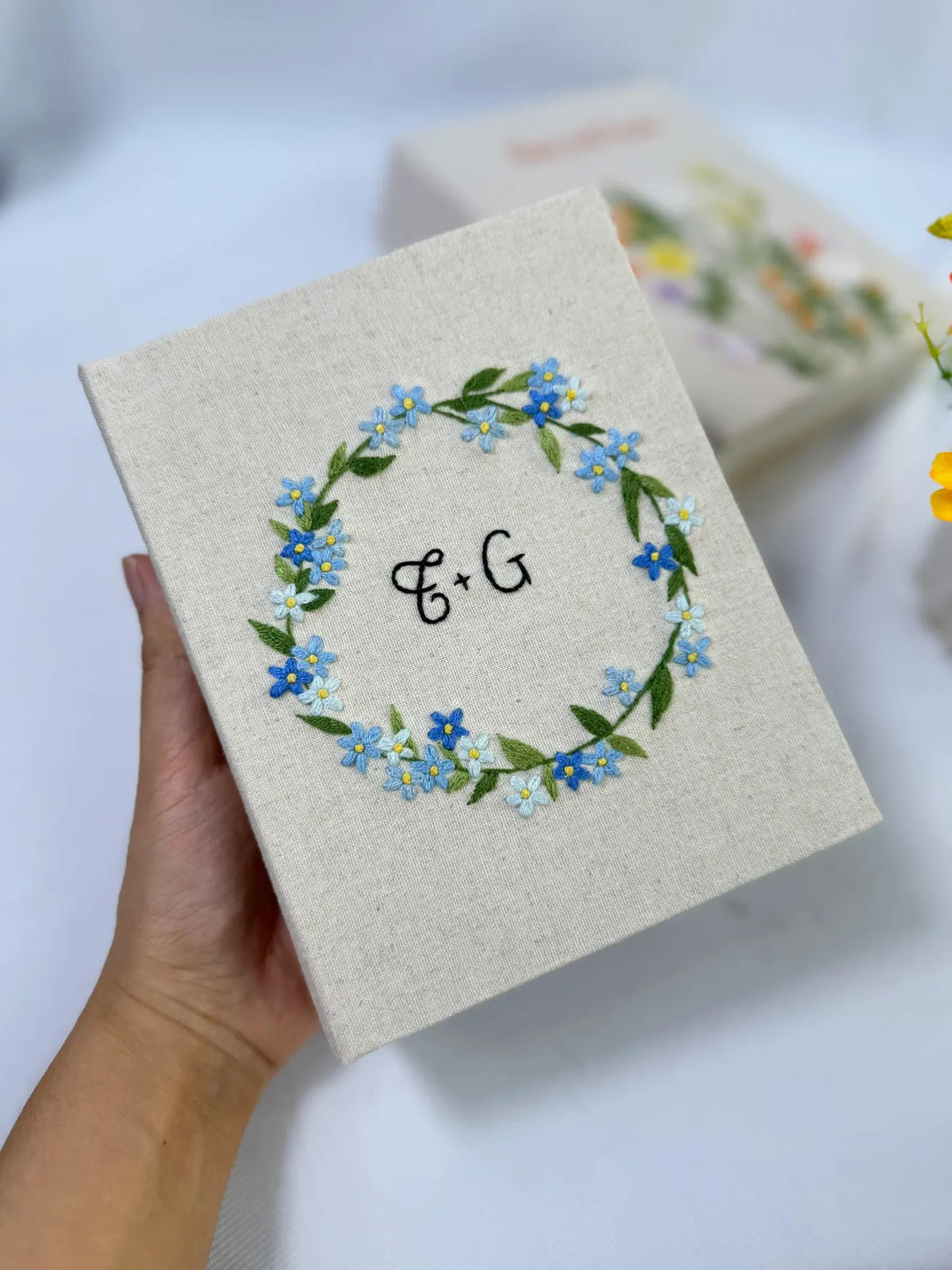 A beige linen book with an embroidered floral wreath in blue and green — hand-embroidered by The Thread Nest