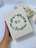 A beige linen book with an embroidered floral wreath in blue and green — hand-embroidered by The Thread Nest