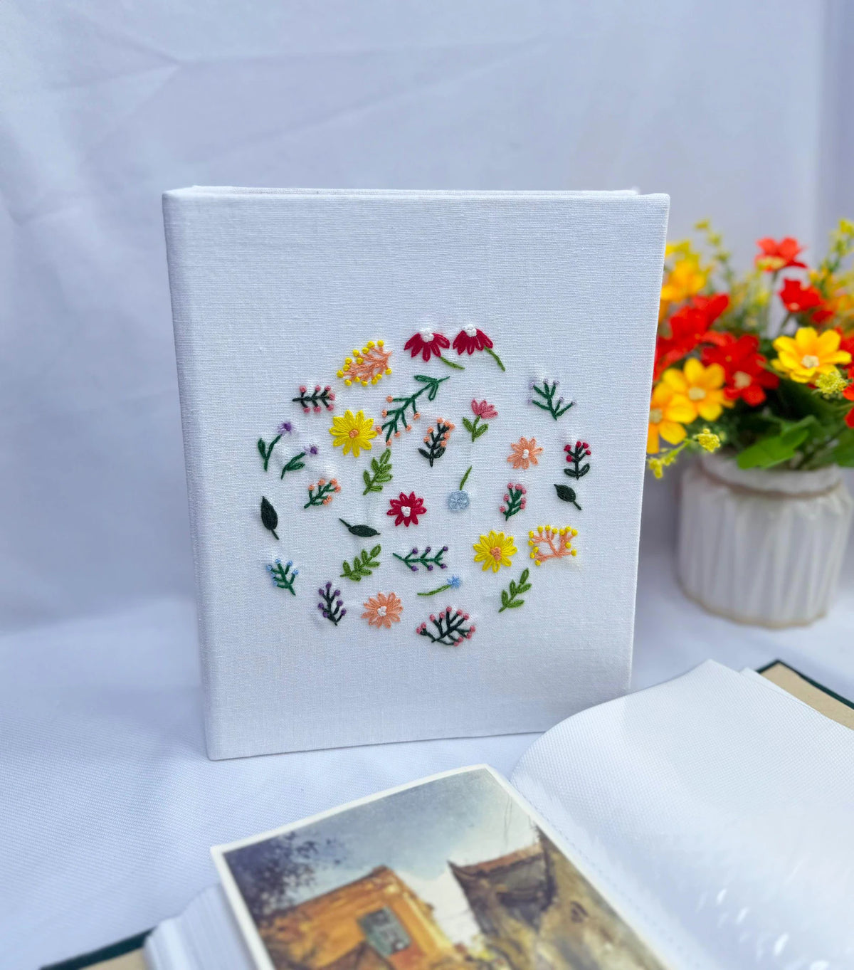 A white photo album with a circular arrangement of embroidered colorful flowers and green leaves — hand-embroidered by The Thread Nest