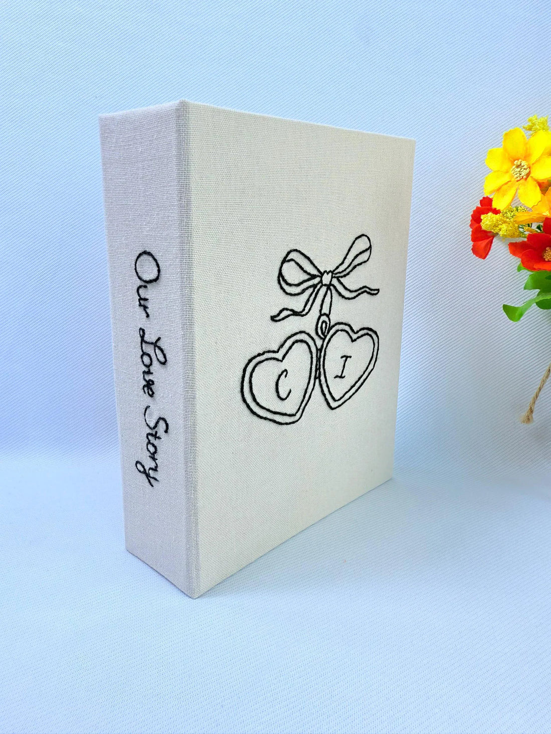 A cream-colored photo album with black embroidered hearts and Our Love Story on the spine — hand-embroidered by The Thread Nest