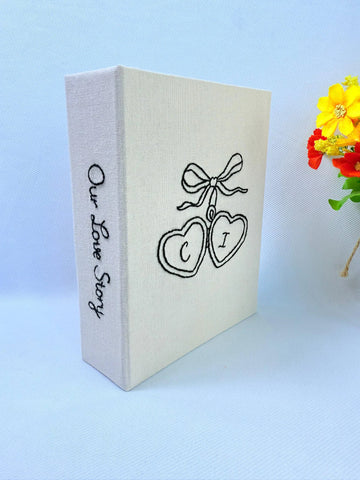 A cream-colored photo album with black embroidered hearts and Our Love Story on the spine — hand-embroidered by The Thread Nest