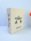 A beige photo album with black embroidered line art and Our Love Story on the spine — hand-embroidered by The Thread Nest