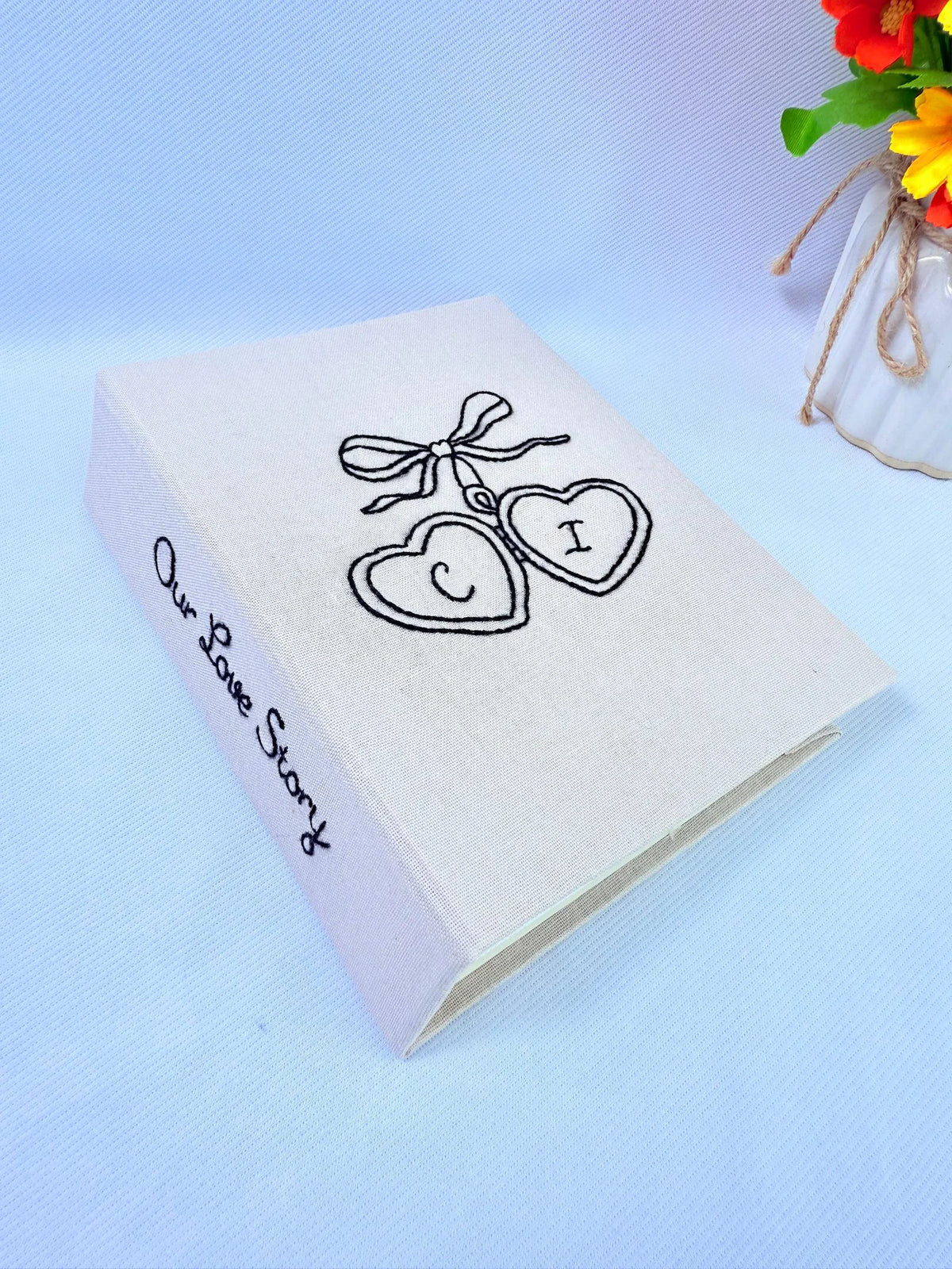 A beige photo album with Our Love Story embroidered in black on the spine — hand-embroidered by The Thread Nest