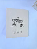 An off-white book with black line art of two clinking champagne glasses — hand-embroidered by The Thread Nest