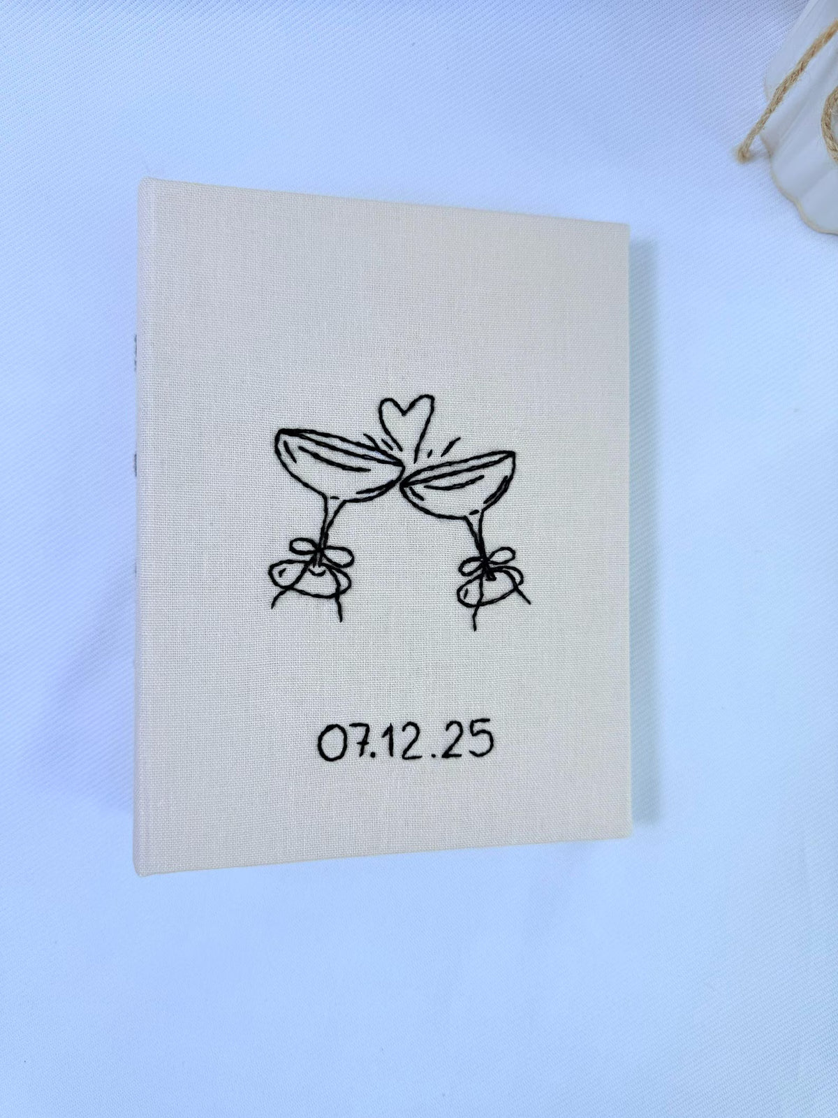 An off-white book with black line art of two clinking champagne glasses — hand-embroidered by The Thread Nest