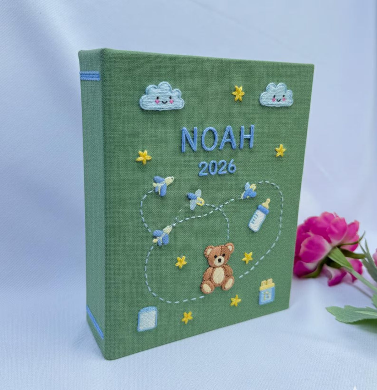 A sage green photo album with a stitched name, a brown teddy bear and clouds — hand-embroidered by The Thread Nest