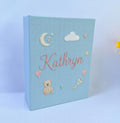 A light blue photo album with an embroidered crescent moon, stars, a teddy bear and a rattle — hand-embroidered by The Thread Nest