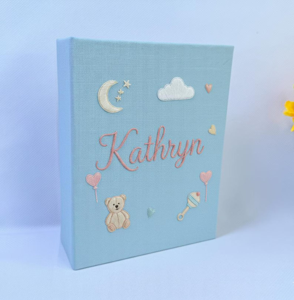 A light blue photo album with an embroidered crescent moon, stars, a teddy bear and a rattle — hand-embroidered by The Thread Nest