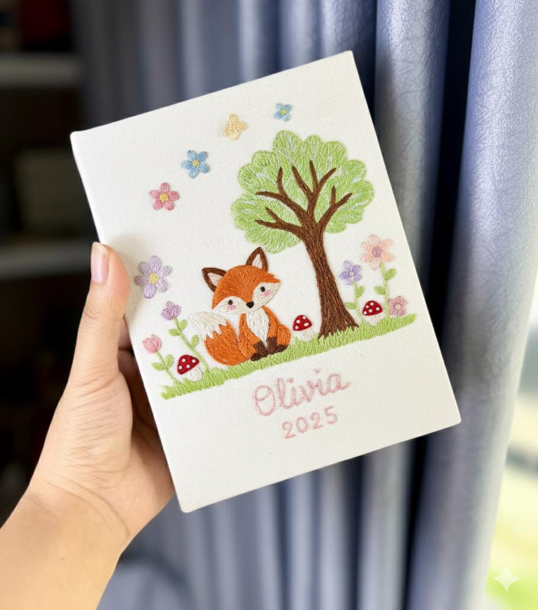 A white notebook with an embroidered fox under a green tree with red mushrooms — hand-embroidered by The Thread Nest