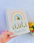 A white keepsake box with an embroidered rainbow, a smiling bee and flowers — hand-embroidered by The Thread Nest