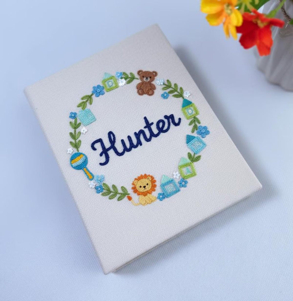 An off-white cover with an embroidered wreath, a teddy bear and a lion — hand-embroidered by The Thread Nest