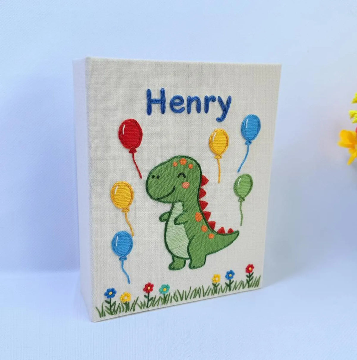A canvas with an embroidered green dinosaur surrounded by colourful balloons — hand-embroidered by The Thread Nest