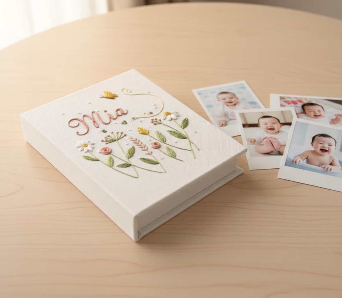 A white photo album with embroidered flowers in green, pink and yellow and the name Mia — hand-embroidered by The Thread Nest