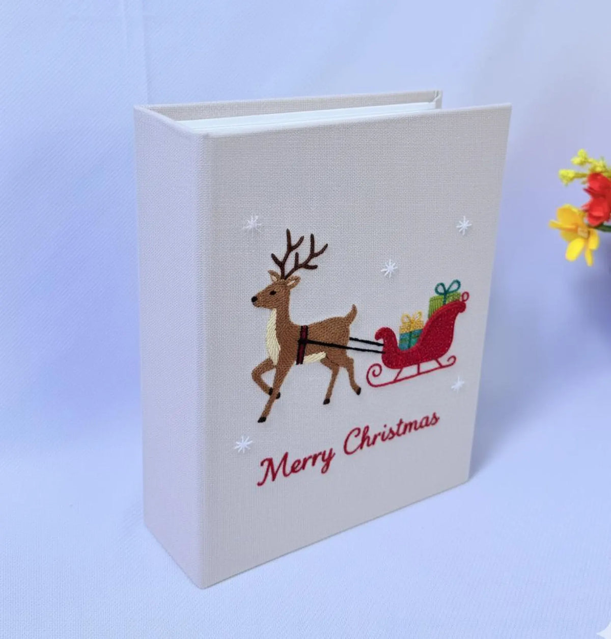 A beige photo album with an embroidered reindeer pulling a red sleigh — hand-embroidered by The Thread Nest