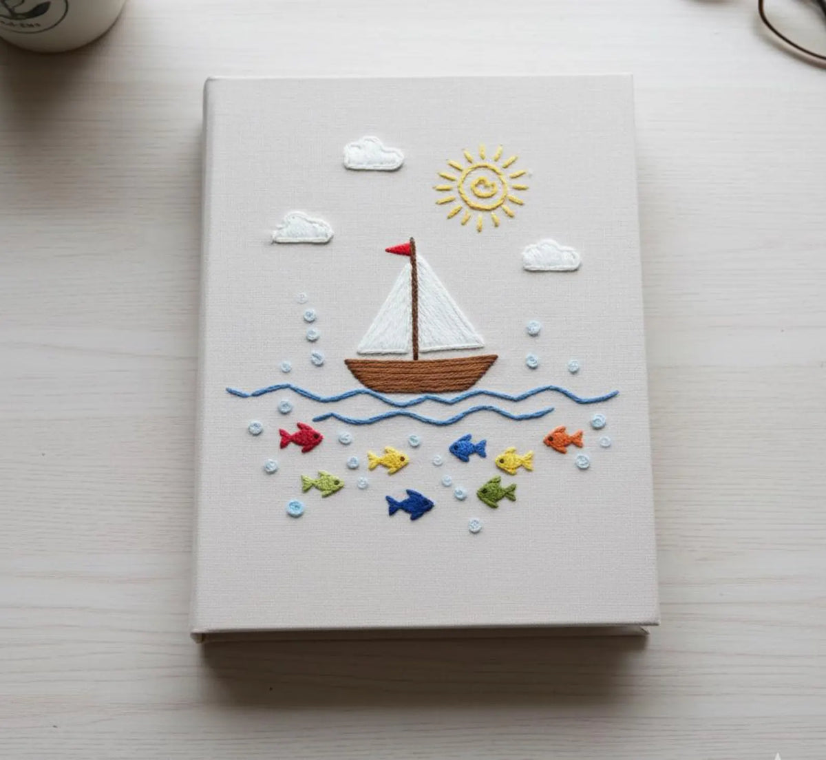 A beige book with an embroidered sailboat, sun, clouds and waves — hand-embroidered by The Thread Nest