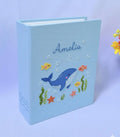 A light blue book with an embroidered blue whale, fish and seaweed — hand-embroidered by The Thread Nest