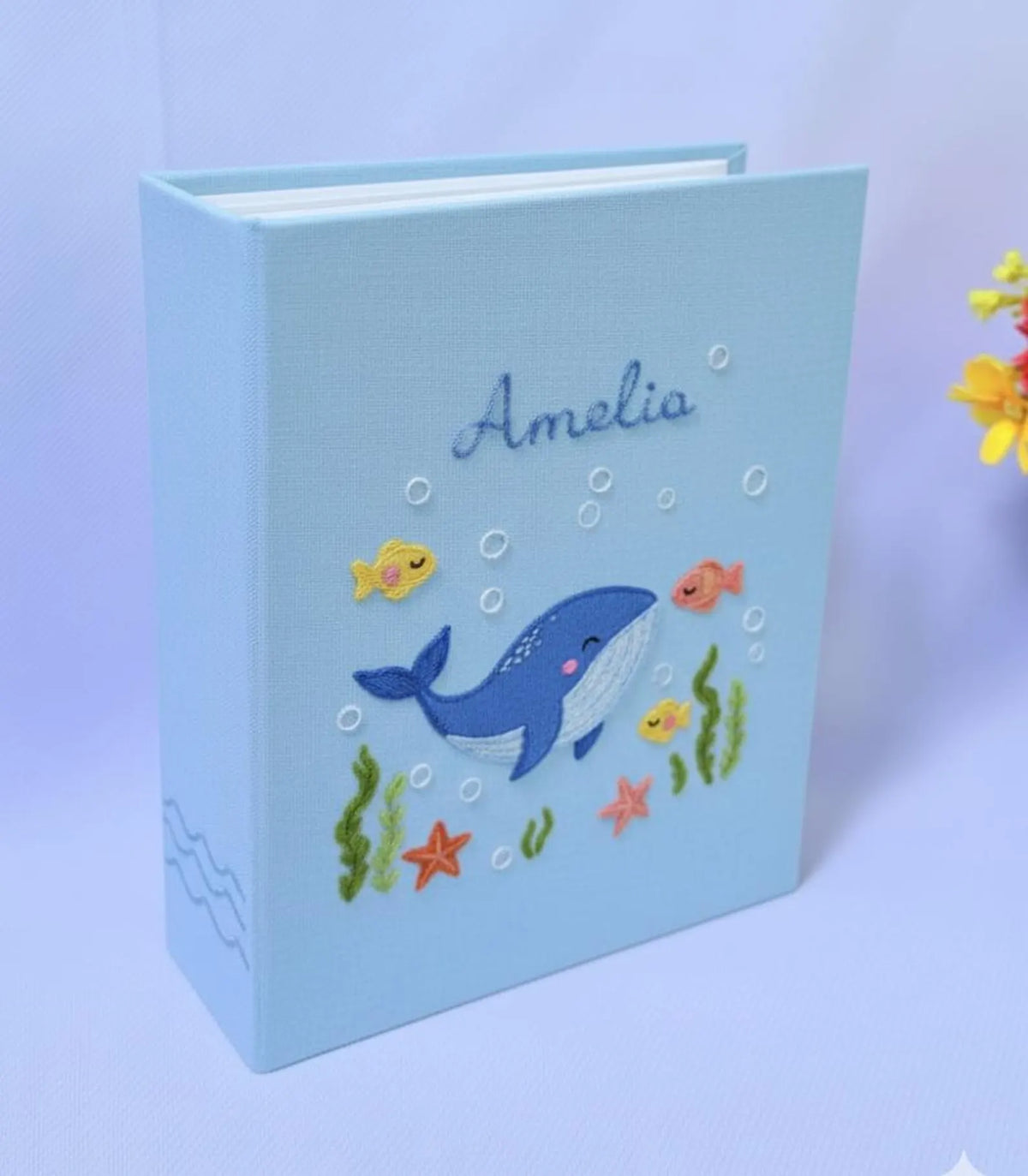 A light blue book with an embroidered blue whale, fish and seaweed — hand-embroidered by The Thread Nest