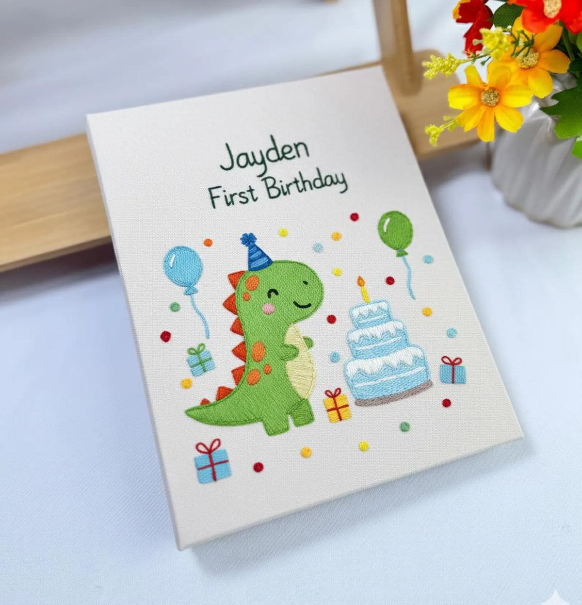 An embroidered canvas with a green dinosaur in a party hat and a birthday cake — hand-embroidered by The Thread Nest