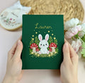 A dark green journal cover with a white bunny, red mushrooms and a name — hand-embroidered by The Thread Nest