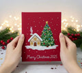 A red Christmas card with an embroidered snow-covered house and tree — hand-embroidered by The Thread Nest