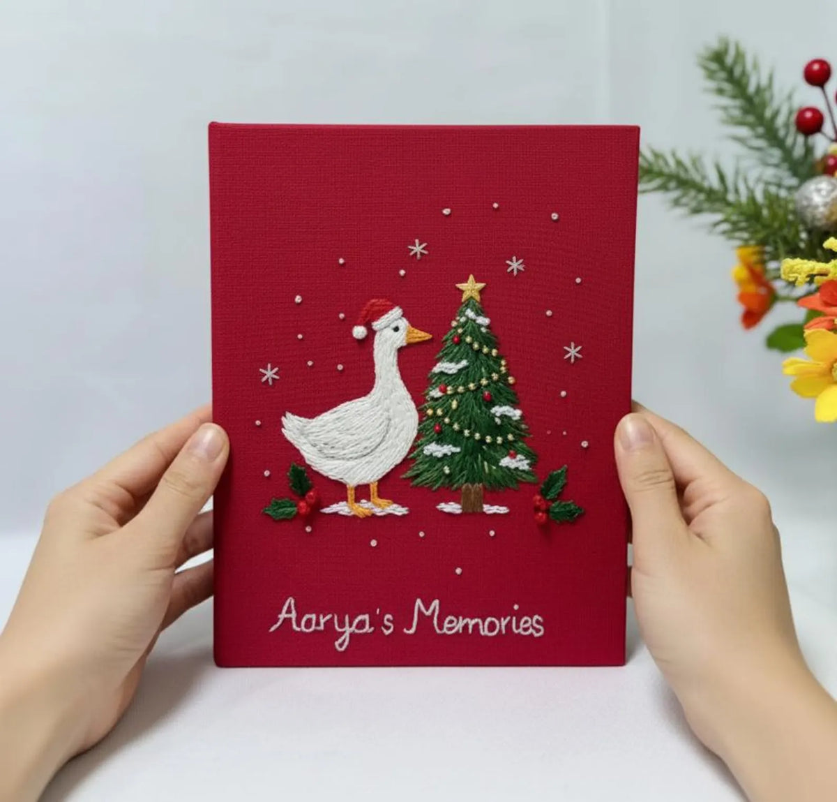 A red photo album with an embroidered goose in a Santa hat beside a Christmas tree — hand-embroidered by The Thread Nest