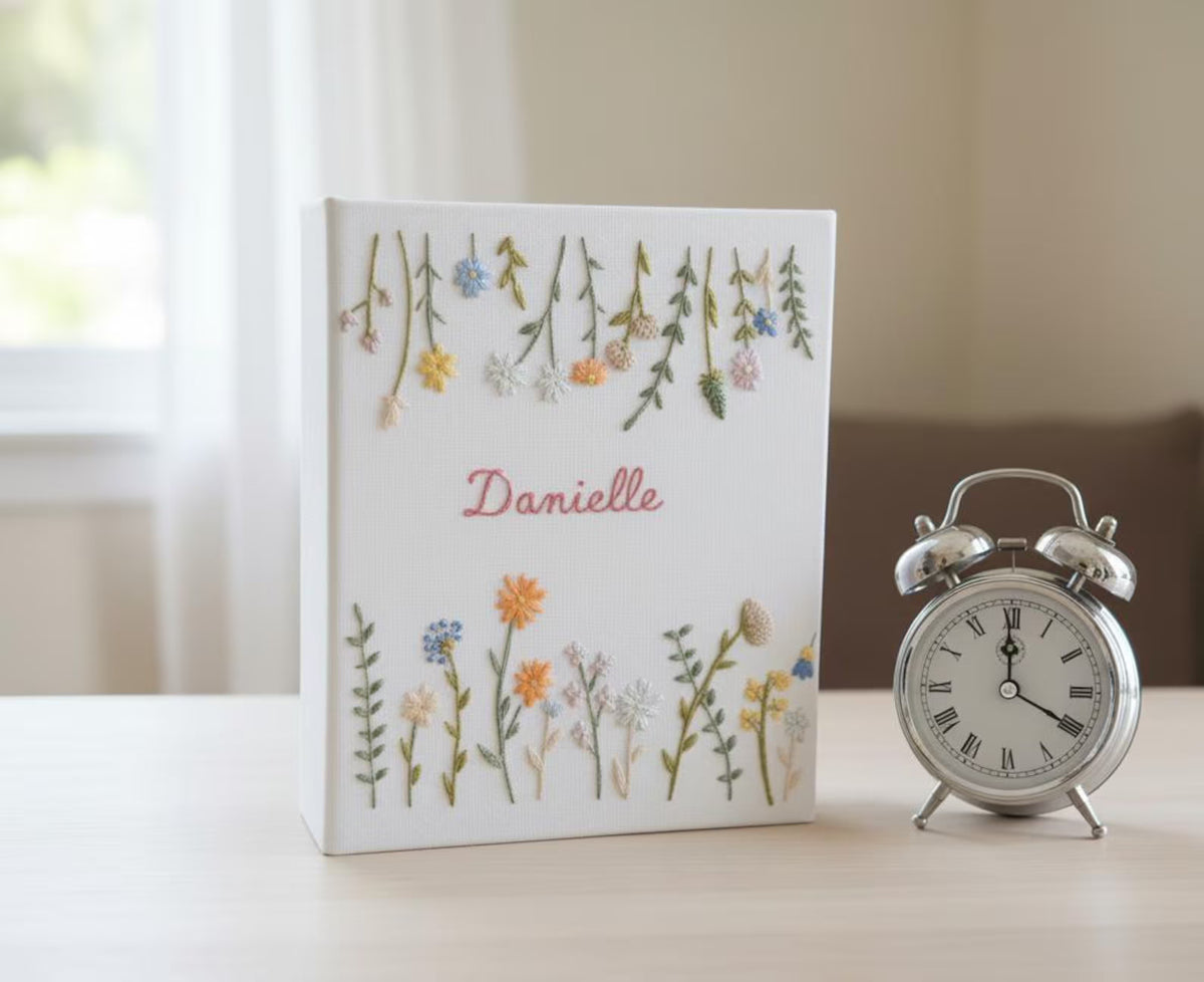 A white book with embroidered flowers and a name in red script — hand-embroidered by The Thread Nest