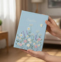 A light blue journal with a stitched name, daisies, roses and lavender — hand-embroidered by The Thread Nest