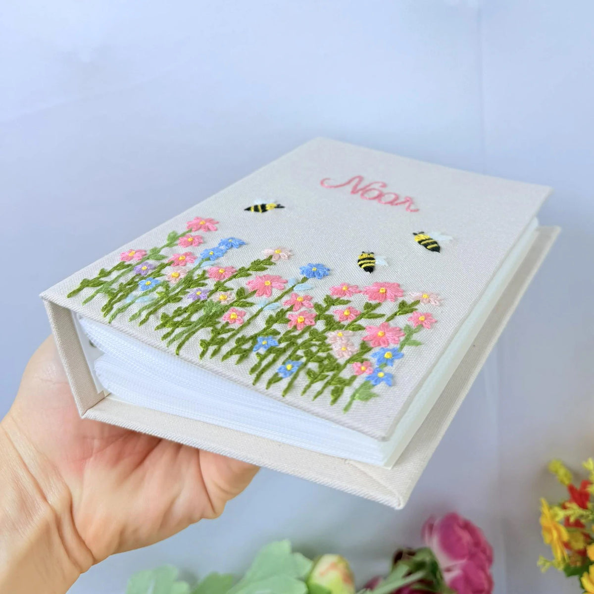 A light beige notebook with hand-embroidered pink and blue wildflowers and bees by The Thread Nest