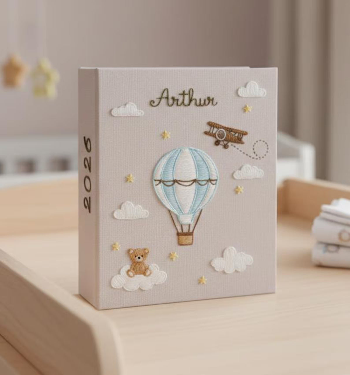 A light beige photo album with an embroidered hot air balloon, airplane and teddy bear — hand-embroidered by The Thread Nest
