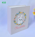 A light pink book with an embroidered white bunny inside a floral wreath — hand-embroidered by The Thread Nest