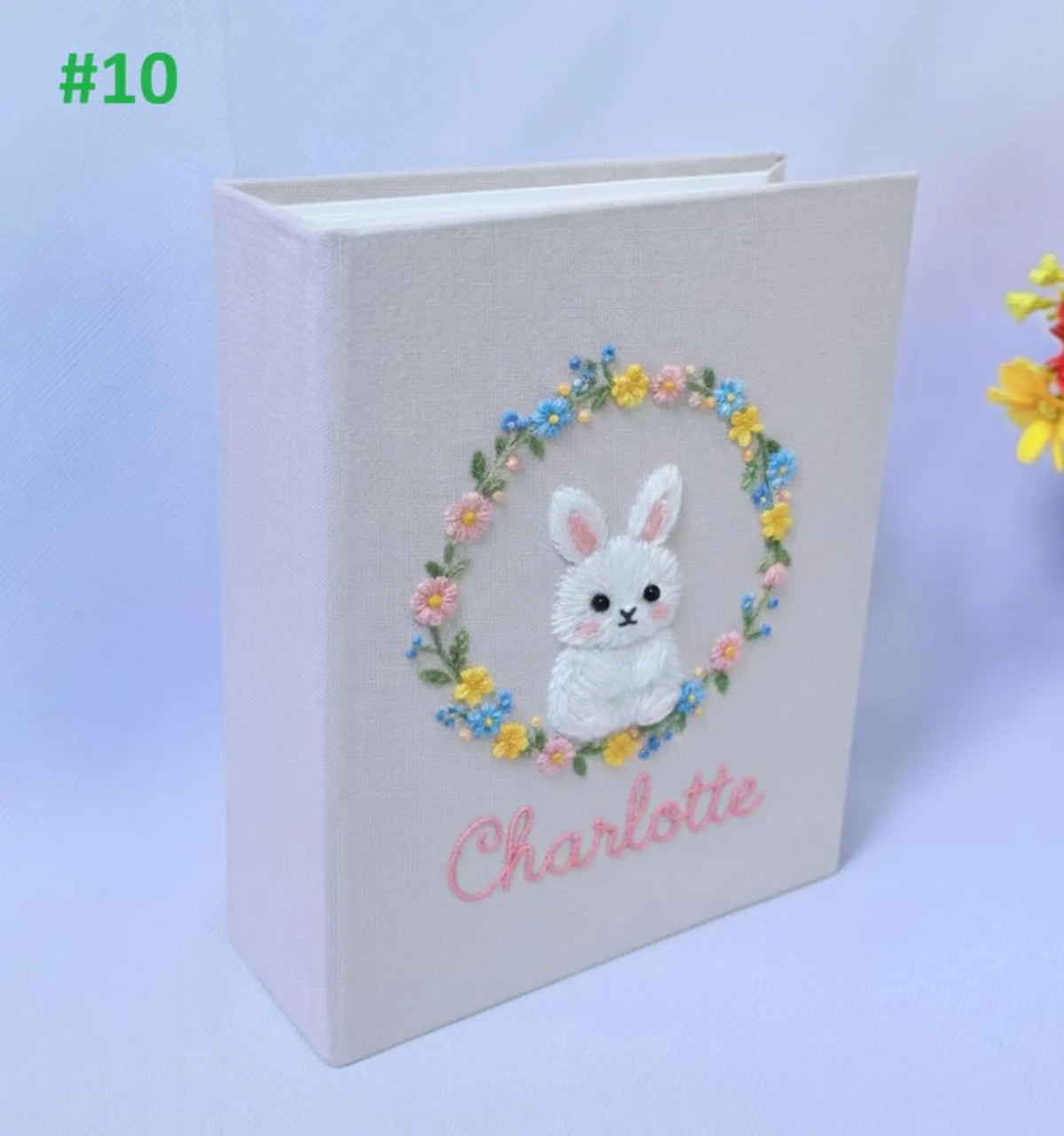 A light pink book with an embroidered white bunny inside a floral wreath — hand-embroidered by The Thread Nest