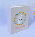 A light pink book with an embroidered white bunny inside a floral wreath — hand-embroidered by The Thread Nest