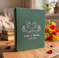A forest green photo album with white embroidered swans facing each other — hand-embroidered by The Thread Nest