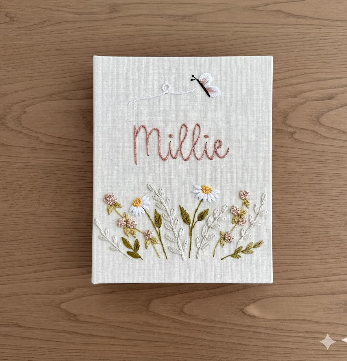 A rectangular white canvas featuring hand-embroidered daisies and the name Millie by The Thread Nest