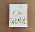 A rectangular white canvas with hand-embroidered daisies and a stitched name by The Thread Nest