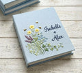 A light blue book with embroidered wildflowers and small bee motifs — hand-embroidered by The Thread Nest