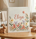 A cream-colored book with a name in peach thread and a surrounding floral design — hand-embroidered by The Thread Nest