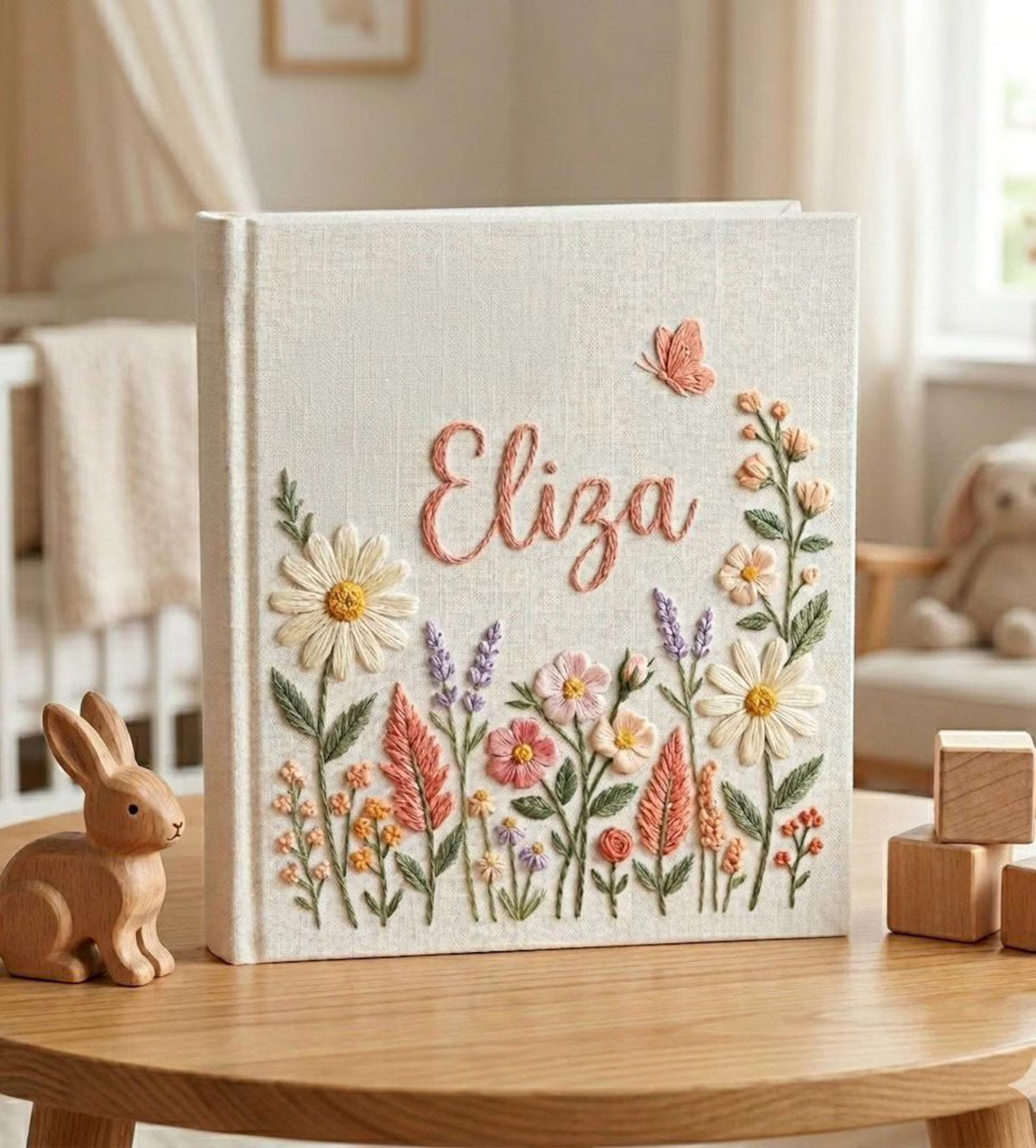 A cream-colored book with a name in peach thread and a surrounding floral design — hand-embroidered by The Thread Nest