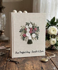 A linen-covered wedding guest book with an embroidered floral bouquet design in white, pink, and green — hand-embroidered by The Thread Nest