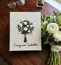 A cream-colored book cover with an embroidered bouquet of white flowers and green leaves — hand-embroidered by The Thread Nest
