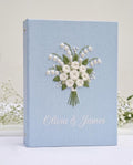 A light blue book with embroidered white roses and lily of the valley — hand-embroidered by The Thread Nest